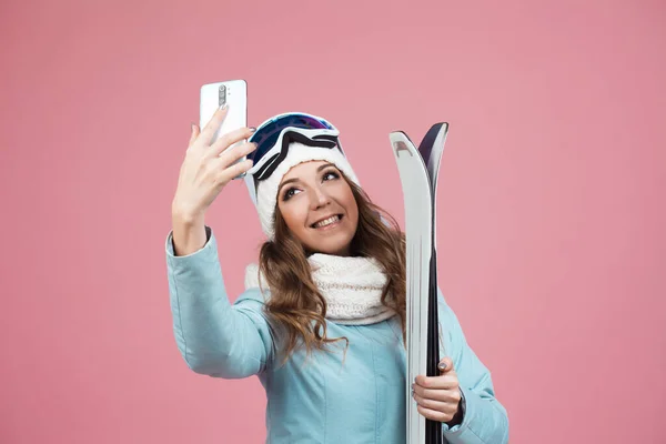 Skieuse amusante prend un selfie sur son smartphone. Une jeune femme avec du matériel de ski en veste et chapeau, — Photo