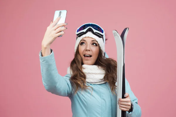 Skieuse amusante prend un selfie sur son smartphone. Une jeune femme avec du matériel de ski en veste et chapeau, — Photo