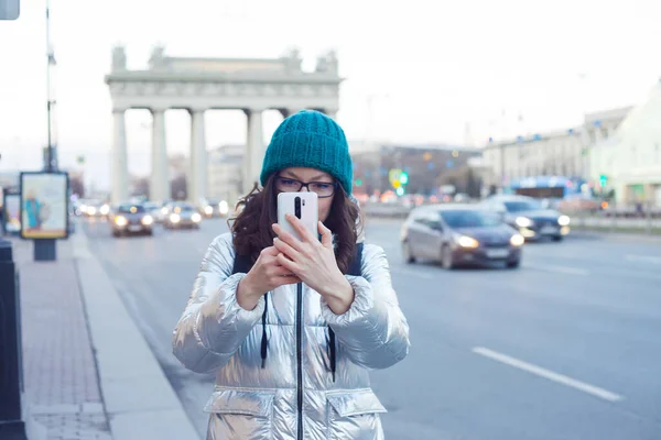 Ein Mädchen in Daunenjacke und Hut läuft durch die Stadt und macht Fotos mit dem Smartphone, — Stockfoto