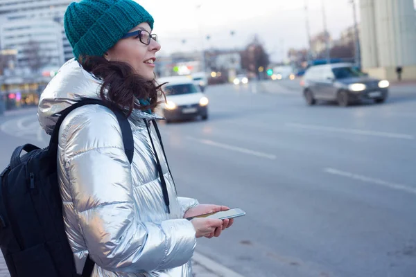 girl in a down jacket and hat stands at a public transport stop and uses a smartphone. Wait for the bus.