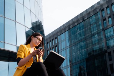 Parlak sarı ceketli genç bir kadın dizüstü bilgisayar ve akıllı telefon kullanıyor..