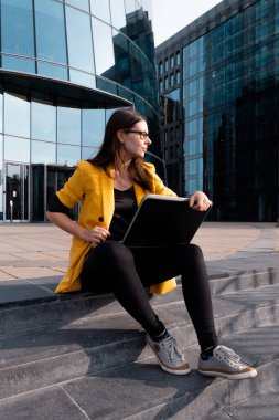 Parlak sarı ceketli genç bir kadın dizüstü bilgisayar ve akıllı telefon kullanıyor..