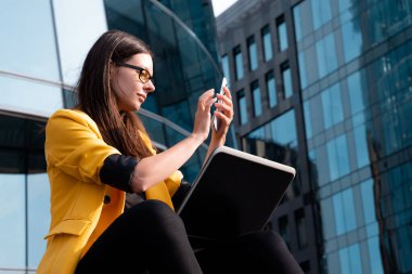 Parlak sarı ceketli genç bir kadın dizüstü bilgisayar ve akıllı telefon kullanıyor..