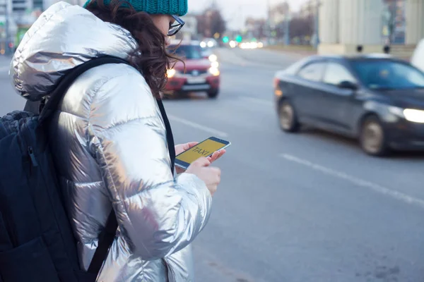 Per Handy-App ein Taxi anrufen, eine junge Frau in Daunenjacke und Hut steht am Straßenrand und wartet auf ein Auto — Stockfoto