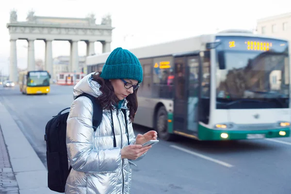 girl in a down jacket and hat stands at a public transport stop and uses a smartphone. Wait for the bus.