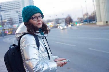 Kapüşonlu ve şapkalı bir kız toplu taşıma durağında duruyor ve akıllı telefon kullanıyor. Otobüsü bekle..