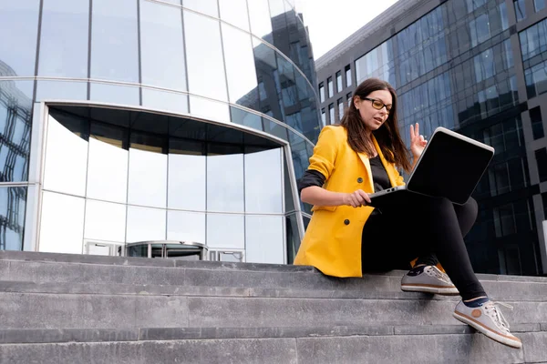 Parlak sarı ceketli genç bir kadın dizüstü bilgisayar ve akıllı telefon kullanıyor..