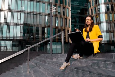 Parlak sarı ceketli genç bir kadın dizüstü bilgisayar ve akıllı telefon kullanıyor..