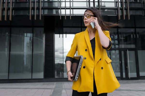 Mujer joven con una chaqueta de color amarillo brillante sostiene un ordenador portátil y el uso de un teléfono inteligente. —  Fotos de Stock