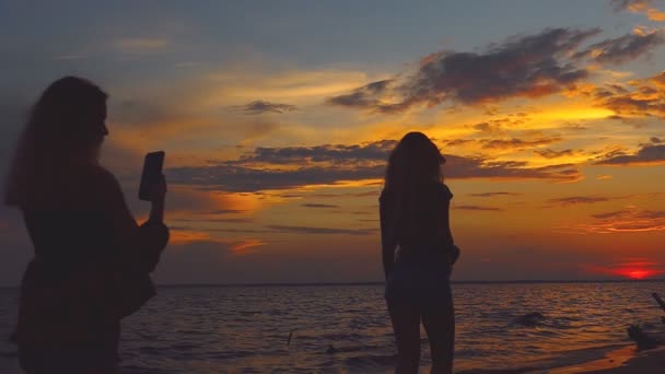 一个年轻的女孩欣赏海滩上的大海景色 并在电话上拍其他女孩的照片 夕阳海滩上轮廓的女人 剪影在日落 — 图库视频影像