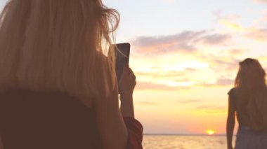 Genç bir kız sahilde deniz görünümü sahiptir ve diğer kız telefonda fotoğraf çeker. Sunset beach kontrast kadın. Gün batımında siluetleri.