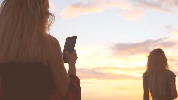 一个年轻的女孩欣赏海滩上的大海景色 并在电话上拍其他女孩的照片 夕阳海滩上轮廓的女人 剪影在日落 — 图库视频影像