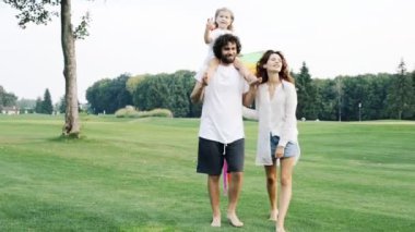 Mutlu bir aile birlikte parkta yürüyüş. Uçurtma tutarak ve babalar omuzlarında oturan çocuk kız.