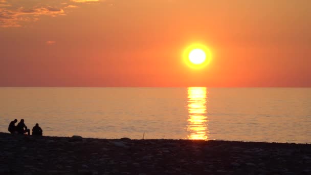 Три Человека Сидят Галечном Пляже Моря Сидят Смотрят Порядок Солнце — стоковое видео