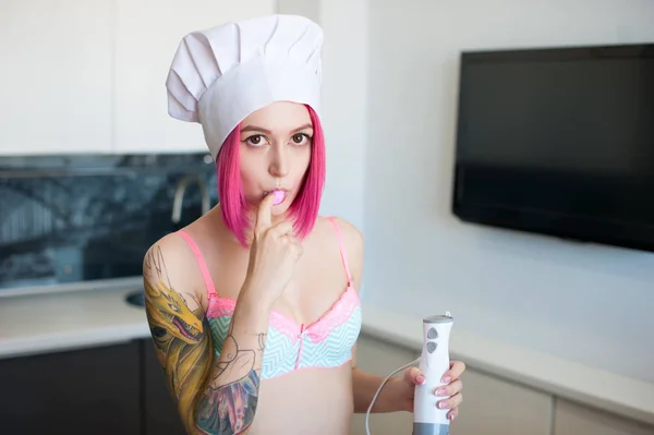 Sexy young emo girl with pink hair in a chef\'s cap tries pink cream in the kitchen
