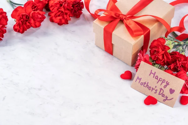 Mayo madres día hecho a mano regalos cuadro desea fotografía - Hermosos claveles en flor con caja de lazo de cinta roja aislado en el escritorio de mármol moderno, primer plano, espacio para copiar, maqueta — Foto de Stock