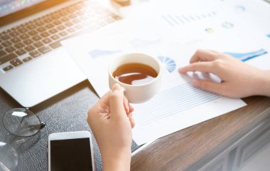 İş konsepti. Kadın raporları okuyor ve ofis masasında dinlenmek için içiyor. Arka aydınlatma, güneş parlama etkisi, yakın, yan görünüm, kopyalama alanı