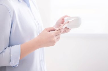 Asya genç iş kadını yan görünümü mavi gömlek pencere yanında duvarda yatan ayakta içme ve akıllı telefon kullanarak, kopya alanı, yakın