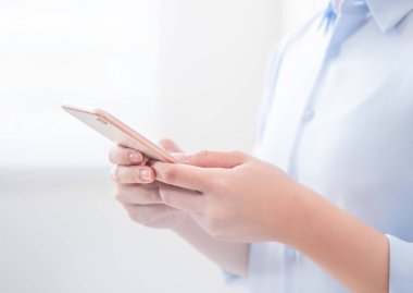 Mavi gömlekli asyalı genç iş kadını yan görünümü pencerenin yanında duran ve parlak ofis odasında akıllı telefon kullanarak, kopya alanı, yakın