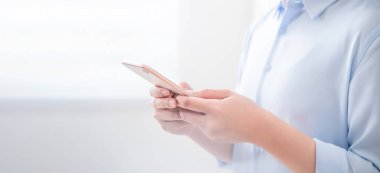 Mavi gömlekli asyalı genç iş kadını yan görünümü pencerenin yanında duran ve parlak ofis odasında akıllı telefon kullanarak, kopya alanı, yakın