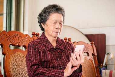 Asyalı yaşlı kadın oturan ve modern akıllı telefon bir şey üzerinden bakarak, evde başkaları ile bağlantı yapma, teknoloji yaşayan, yakın