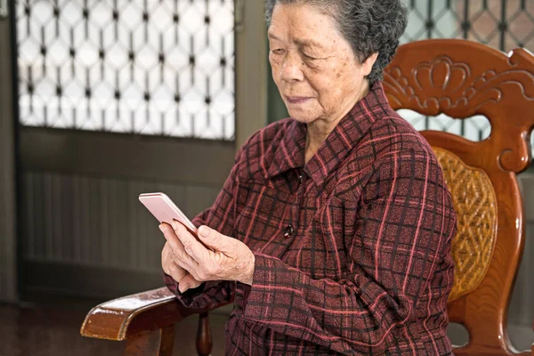 Asian elderly woman sitting and looking through something on modern smartphone, making connection with others at home, living technology, close up