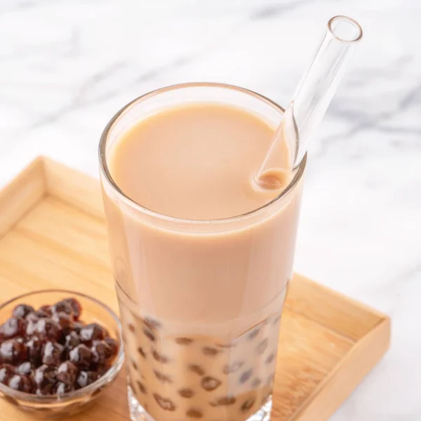 Tapioca pearl ball bubble milk tea, popular Taiwan drink, in drinking glass with straw on marble white table and wooden tray, close up, copy space.