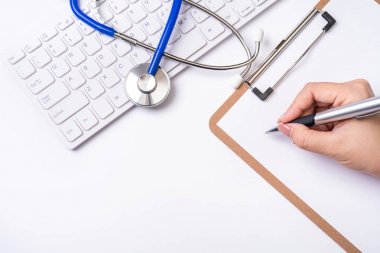 Kadın doktor steteskop, bilgisayar klavyesi ile beyaz çalışma masası üzerinde pano üzerinde tıbbi kayıt durumda yazıyor. Üst görünüm, düz döşeme, kopyalama alanı
