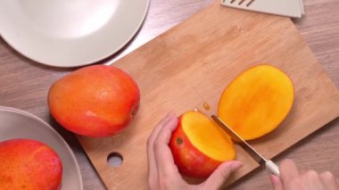 Genç kadın ahşap bir masada yemek için güzel bir taze sulu mango kesiyor ve mutfakta doğrama tahtası, yakın , 4k video çekim.