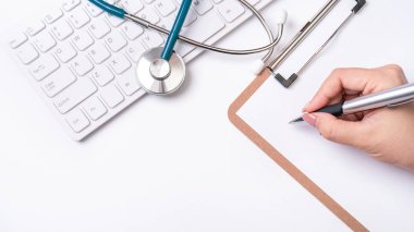 Kadın doktor steteskop, bilgisayar klavyesi ile beyaz çalışma masası üzerinde pano üzerinde tıbbi kayıt durumda yazıyor. Üst görünüm, düz döşeme, kopyalama alanı