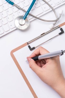 Kadın doktor steteskop, bilgisayar klavyesi ile beyaz çalışma masası üzerinde pano üzerinde tıbbi kayıt durumda yazıyor. Üst görünüm, düz döşeme, kopyalama alanı