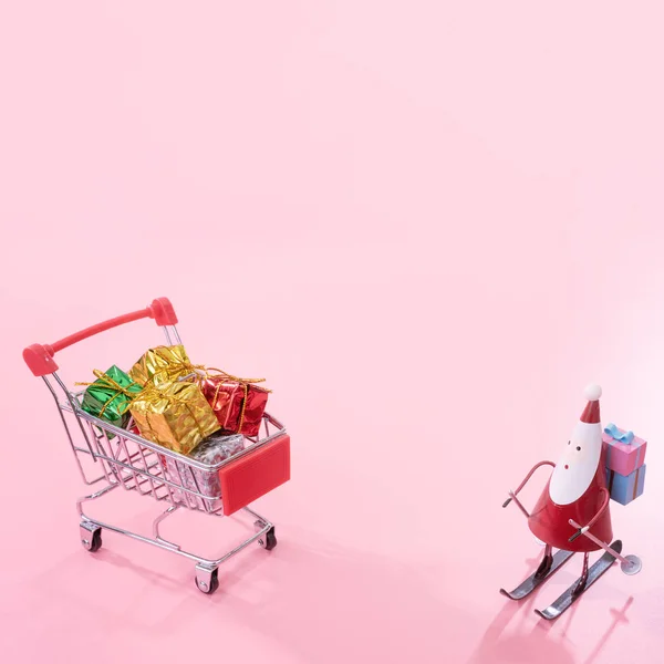 Karácsonyi bevásárló koncepció, mini piros üzlet cart kocsi, Mikulás játék és ajándékdoboz izolált halvány rózsaszín háttér, üres másolási tér, közelről — Stock Fotó