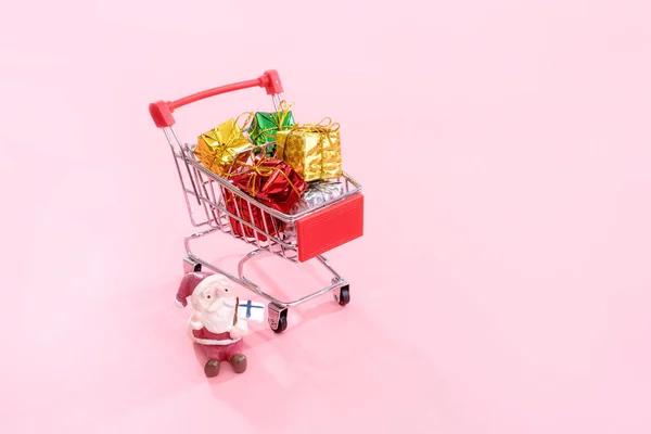 Conceito de compras de Natal, mini carrinho de compras vermelho carrinho com brinquedo Papai Noel e caixa de presente isolado em fundo rosa pálido, espaço em branco cópia, close-up — Fotografia de Stock