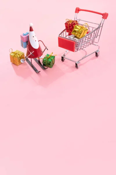 Karácsonyi bevásárló koncepció, mini piros üzlet cart kocsi, Mikulás játék és ajándékdoboz izolált halvány rózsaszín háttér, üres másolási tér, közelről — Stock Fotó
