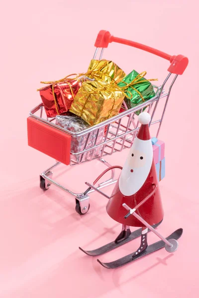 Kerst winkelen concept, mini rode winkel kar trolley met Santa Claus speelgoed en geschenk doos geïsoleerd op bleke roze achtergrond, lege kopie ruimte, close-up — Stockfoto