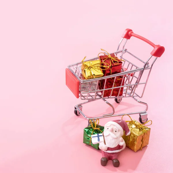 Kerst winkelen concept, mini rode winkel kar trolley met Santa Claus speelgoed en geschenk doos geïsoleerd op bleke roze achtergrond, lege kopie ruimte, close-up — Stockfoto