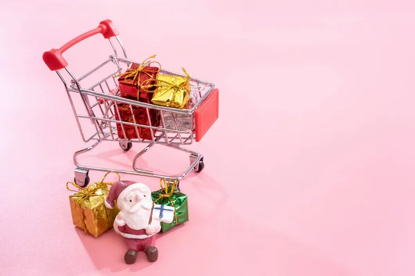 Kerst winkelen concept, mini rode winkel kar trolley met Santa Claus speelgoed en geschenk doos geïsoleerd op bleke roze achtergrond, lege kopie ruimte, close-up — Stockfoto