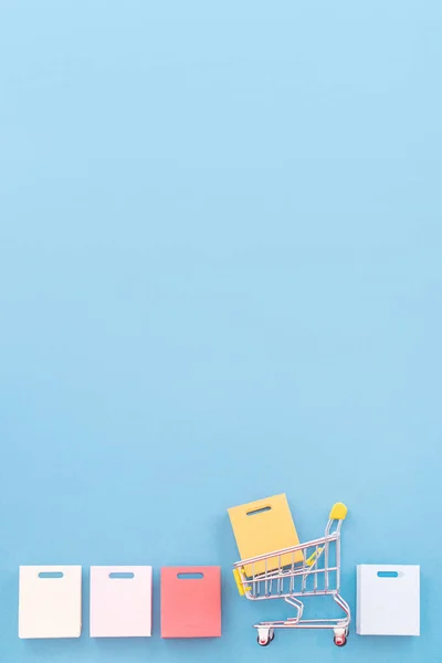 Abstraktes Gestaltungselement, Jahresverkauf, Konzept der Einkaufssaison, kleiner gelber Einkaufswagen mit bunter Papiertüte auf pastellblauem Hintergrund, Draufsicht, flache Lage — Stockfoto