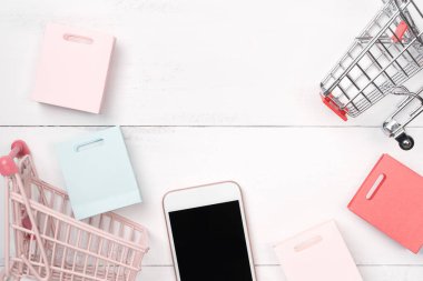 Soyut online alışveriş, mobil ödeme konsept tasarım unsuru, beyaz ahşap masa zemininde renkli el arabası ve kağıt torbalar, üst görünüm, düz yatış