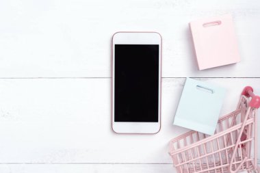 Soyut online alışveriş, mobil ödeme konsept tasarım unsuru, beyaz ahşap masa zemininde renkli el arabası ve kağıt torbalar, üst görünüm, düz yatış