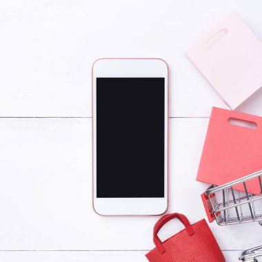 Soyut online alışveriş, mobil ödeme konsept tasarım unsuru, beyaz ahşap masa zemininde renkli el arabası ve kağıt torbalar, üst görünüm, düz yatış