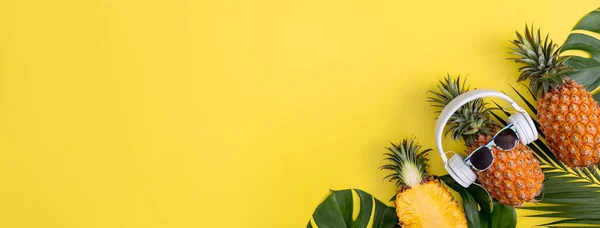 Funny pineapple wearing white headphone, listen music, isolated on yellow background with tropical palm leaves, top view, flat lay design concept.