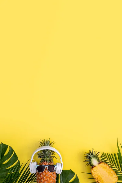 Funny pineapple wearing white headphone, listen music, isolated on yellow background with tropical palm leaves, top view, flat lay design concept.
