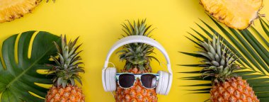 Beyaz kulaklık takan komik ananas, müzik dinle, sarı arka planda tropikal palmiye yapraklarıyla izole edilmiş, üst görünüm, düz dizayn konsepti.
