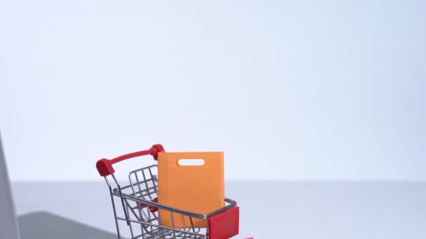 Online shopping. Mini shop cart trolley with colorful paper bags and laptop computer on white table background, buying at home concept, close up