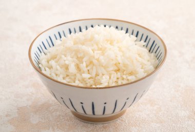 Bir kase taze pişmiş beyaz pirinç, yumuşak, kabarık ve beyaz masa arkasında hafifçe parıltılı..