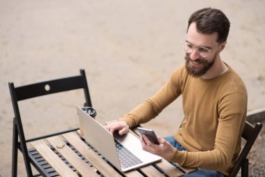 yakışıklı sakallı adam dizüstü bilgisayarda çalışan ve ahşap masada akıllı telefon tutarak