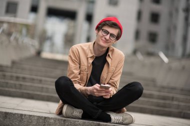 genç genç çocuk, bir hipster, kırmızı bir şapka ve şık bir gömlek metropol şehir yürür ve müzik dinlerken ve iyi bir ruh hali ile dinlenmek olan telefonunda bir şey bakar