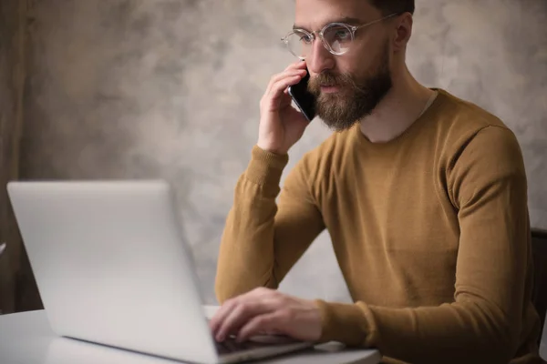 Przystojny Brodaty Mężczyzna Okularach Pracujących Laptopie Kawiarni Rozmawiając Przez Smartphone — Zdjęcie stockowe