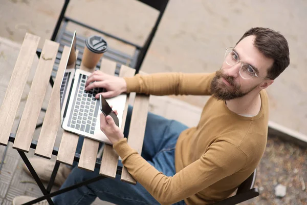 handsome bearded man working at laptop and holding smartphone on wooden table